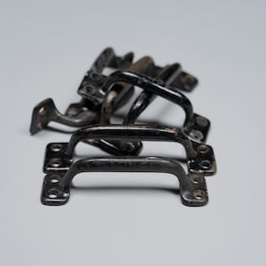 May include: A collection of black metal handles with a curved grip and rectangular mounting plates. Each plate has multiple screw holes. The handles appear to be vintage or antique, with signs of wear and tear.