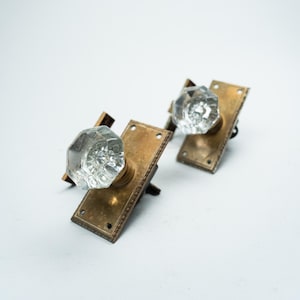 Peut inclure: Deux poignées de porte anciennes avec des boutons en verre clair à facettes et des plaques arrière rectangulaires en laiton. La quincaillerie de porte est posée sur un fond blanc. Les boutons ont un design classique et vintage.