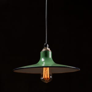 May include: A green pendant light with a black cord and a vintage-style lightbulb. The light fixture has a wide, shallow shade with a white interior and a black trim. The light is suspended against a black background.