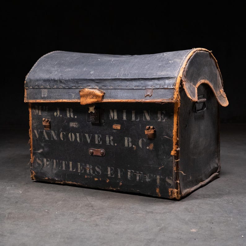 May include: An antique black trunk with a curved top and leather accents. The trunk has the text "J.R. MILNE VANCOUVER R.B.C. SETTLERS EFFECTS" printed on the front. The trunk is made of a dark material and has metal clasps and leather handles.