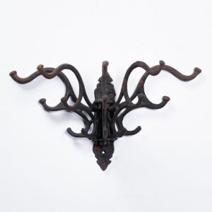 Puede incluir: Un perchero de hierro fundido de color marrón oscuro con múltiples ganchos. El perchero tiene un diseño ornamentado con brazos y ganchos curvos, ideal para un pasillo. El perchero está montado en una pared blanca.