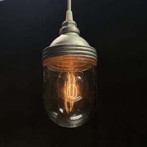 May include: A vintage-style pendant light fixture with a clear glass shade and a metal cap. The exposed filament bulb glows with a warm, orange light. The fixture hangs from a white cord against a dark background.