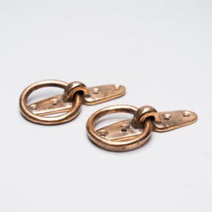 May include: Two bronze-coloured metal D-rings with attached rectangular plates. Each ring is circular and connected to a plate with three holes for mounting. The hardware has a vintage or antique appearance.