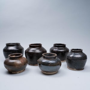 Peut inclure: Ensemble de sept petits pots ronds en céramique, de couleurs marron et noir. Les pots ont un aspect rustique et vieilli, avec des imperfections visibles et une finition mate. Les pots sont de tailles différentes.
