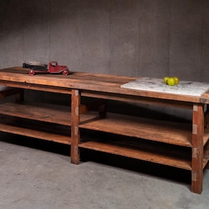 May include: A long, rectangular wooden workbench featuring a natural brown finish, exposed joinery, and two lower shelves. A rectangular white marble slab is inset into the right side of the top surface, with three green apples and a red toy truck.