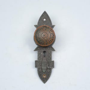 May include: An antique door knob and plate set. The knob is round with a copper finish and ornate floral and geometric patterns. The plate is a dark gray metal with a decorative design and keyhole.