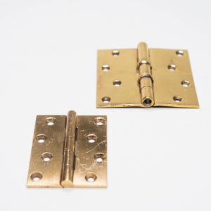 May include: Two brass door hinges, one square and one rectangular, are displayed against a white background. Each hinge has multiple screw holes for mounting. The hinges are made of a shiny, golden-colored metal.
