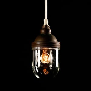 May include: A vintage-style pendant light fixture with a clear glass shade and a bronze-colored metal cap. The light bulb inside is illuminated, casting a warm glow. The fixture hangs from a white cord against a black background.