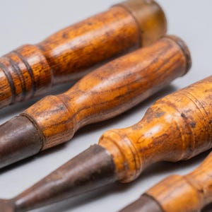 May include: Close-up of several antique wood chisels. The chisels have wooden handles with a warm, brown finish and aged metal blades. The tools are arranged at a slight angle, showcasing their craftsmanship and vintage aesthetic.