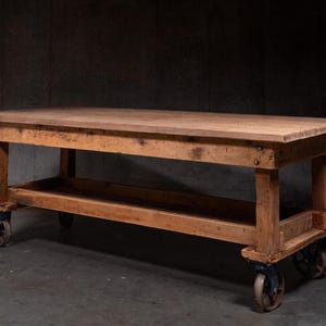 May include: A large, rectangular wooden work table with a natural finish. The table has a lower shelf and is mounted on four antique metal caster wheels. The table is approximately 183 cm long, 91 cm wide, and 91 cm high.