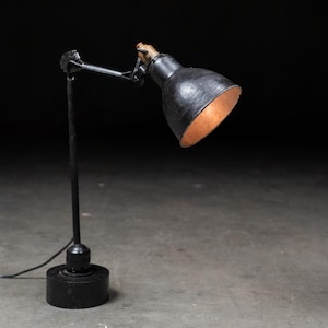 May include: A black desk lamp with an adjustable arm and a round base. The lamp shade is black with a copper-colored interior. The lamp is designed to direct light.
