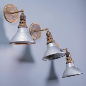 Peut inclure: Trois appliques murales composées de plaques de fixation circulaires en laiton, de bras articulés en laiton et d'abat-jours coniques en métal argenté strié. Chaque lampe possède une douille en laiton ventilée reliant le bras à l'abat-jour.