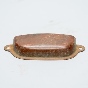 Puede incluir: Un tirador de cajón rectangular de metal con acabado marrón. El tirador tiene una parte superior ligeramente curvada y pequeños orificios circulares en cada extremo para el montaje. El metal muestra signos de edad y desgaste.