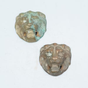 May include: Two metallic lion head ornaments featuring a weathered bronze finish with visible green oxidation. Each piece displays a sculpted mane, defined facial features, and an open mouth design, showcasing a textured and aged appearance.