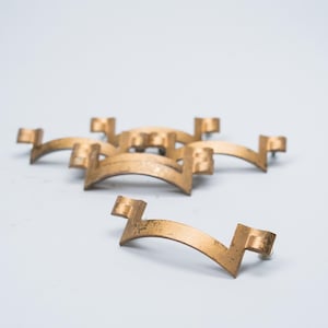 May include: A collection of antique brass-toned cabinet pulls. Each pull features a curved, arched design with rectangular end pieces. The pulls have a slightly distressed finish, suggesting age. They are arranged on a white surface.