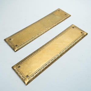 May include: Two rectangular brass door plates. Each plate has a central, smooth section surrounded by a decorative border. The borders feature a repeating, curved design. Each plate has four screw holes for mounting.