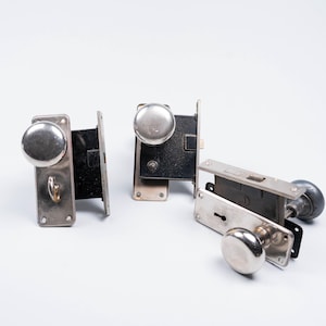 May include: Four antique door lock sets. Each set features a metal plate, a keyhole, and a round doorknob. The doorknobs are silver and the metal plates are silver and black. The locks are displayed on a white surface.