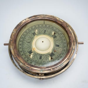 May include: A vintage brass compass with a glass cover. The compass has a circular design with directional markings and degree indicators. The brass casing shows signs of age and wear, adding to its antique aesthetic. The compass is a navigational tool.