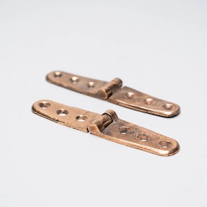 May include: Two antique bronze hinges with rounded edges and multiple screw holes. The hinges are slightly open, showing the joint. The background is a plain white surface.