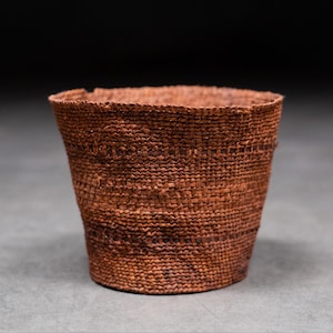 May include: A hand-woven, reddish-brown basket with a slightly flared shape. The basket is made of tightly woven natural fibres, showcasing a textured surface and a rustic aesthetic. The open top and sturdy construction suggest a functional storage item.