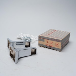 May include: Two chrome towel bar brackets and a small box labeled "Lawson Pilgrim Towel Bar Brackets". A small bag of screws is also present. The brackets are shiny and appear unused.