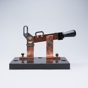 Puede incluir: Un interruptor de cuchilla eléctrico vintage con un mango negro y una cuchilla de cobre. El interruptor está montado sobre una base negra con terminales y pernos de cobre. El interruptor está diseñado para abrir y cerrar un circuito eléctrico.