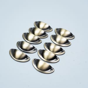 May include: A collection of ten semi-circular metal cup drawer pulls featuring a brushed antique brass finish. Each pull has a curved, recessed handle design with two small screw holes on the flat mounting tabs at either end for attachment.