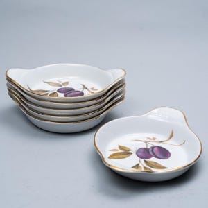 May include: A collection of six white ceramic dishes with gold rims and handles. Each dish displays a purple plum design with green leaves. The dishes are stacked, with one separate, on a grey surface.