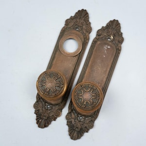 Könnte beinhalten: Zwei antike Türgriffgarnituren aus Bronze. Jedes Set enthält eine lange, rechteckige Rückplatte mit aufwendigen floralen Details und einen runden Türknauf. Die Türknäufe haben ein detailliertes, kreisförmiges Design. Die Sets haben eine warme, gealterte Bronzefarbe.