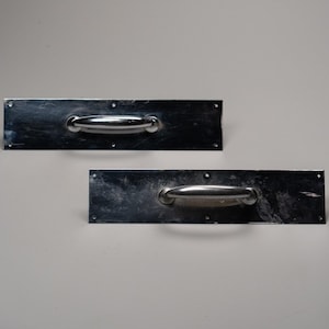 Może przedstawiać: Dwa prostokątne, srebrzyste uchwyty do drzwi z zaokrąglonymi uchwytami. Każdy uchwyt jest zamontowany na prostokątnej płycie z kilkoma otworami na śruby. Uchwyty są ustawione na gładkim, jasnym tle.