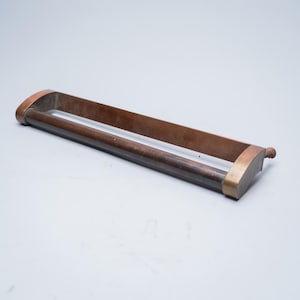 May include: A rectangular, antique-style towel bar with a dark brown wooden handle and copper-colored metal accents. The bar has a simple, minimalist design, with a rounded end and a rectangular frame.