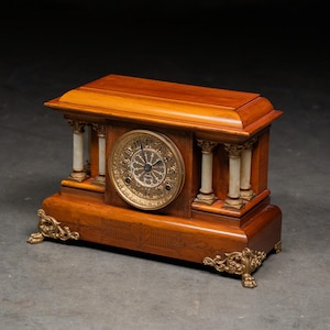 May include: A wooden antique clock with a light brown finish. The clock features a round, gold-colored face with Roman numerals and decorative detailing. It has white columns and gold accents, with ornate feet.