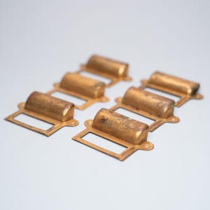 以下が含まれることがあります： ラベルホルダー付きの、真鍮製のアンティーク引き出し取っ手6個。取っ手は長方形のフレームと、湾曲した円筒形のハンドルを備えています。真鍮は風化し、古びた外観で、色と質感にばらつきがあります。