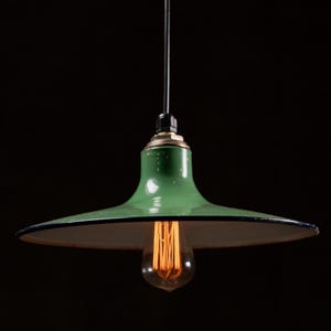 Puede incluir: Lámpara colgante verde de estilo vintage con una pantalla ancha orientada hacia abajo. La pantalla tiene un borde negro y un interior blanco. Una bombilla Edison visible cuelga del centro, emitiendo un brillo cálido. La lámpara está suspendida de un cable negro sobre un fondo oscuro.