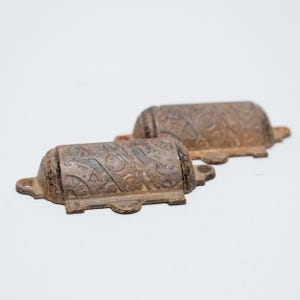 Peut inclure: Deux poignées de tiroir antiques en fonte, avec une finition marron vieillie. Chaque poignée a un corps cylindrique avec des motifs en relief ornés et une base rectangulaire avec des trous de montage. Style vintage ou rustique.