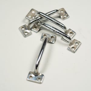 May include: A collection of six shiny, silver-colored metal handles. Each handle has a curved bar and two rectangular mounting plates with two holes each. The handles are likely for cabinets, drawers, or other furniture.