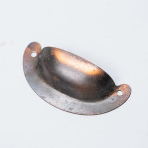 Op de afbeelding: Een verweerde, antieke komgreep in een bronzen afwerking. Het metalen beslag heeft een gebogen ontwerp met twee montagegaten aan beide uiteinden. De binnenkant van de kom heeft een donkerdere patina. Ideaal voor kast- of laderestauratie.