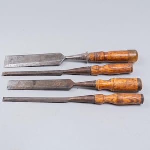 May include: Four antique wood chisels with metal blades and wooden handles. The chisels vary in size and blade shape, showing signs of wear. The handles are a light brown color, and the blades are a darker, aged metal.