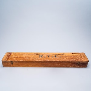 May include: A rectangular wooden box with a hinged lid. The box is a light brown color with visible wood grain and wear. The text "R.T.E." is written on the top in black. The box has small metal clasps.