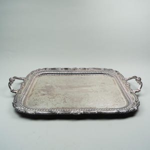 May include: A rectangular silver-toned serving tray featuring ornate, raised edges with a decorative beaded and scrollwork border. The tray includes two curved handles on opposite sides and an engraved floral pattern across the central surface.