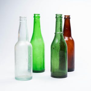 May include: Four glass bottles of different colours and shapes stand against a white backdrop. The bottles include clear, green, and brown hues, showcasing different designs and sizes. The bottles are empty.
