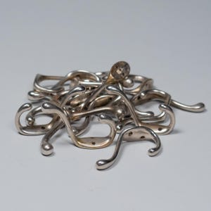 Op de afbeelding: Een stapel zilverkleurige metalen kapstokken. Elke haak heeft een gebogen ontwerp met een afgerond uiteinde en een platte basis met twee gaten voor montage. De haken liggen verspreid op een lichtgrijze ondergrond.