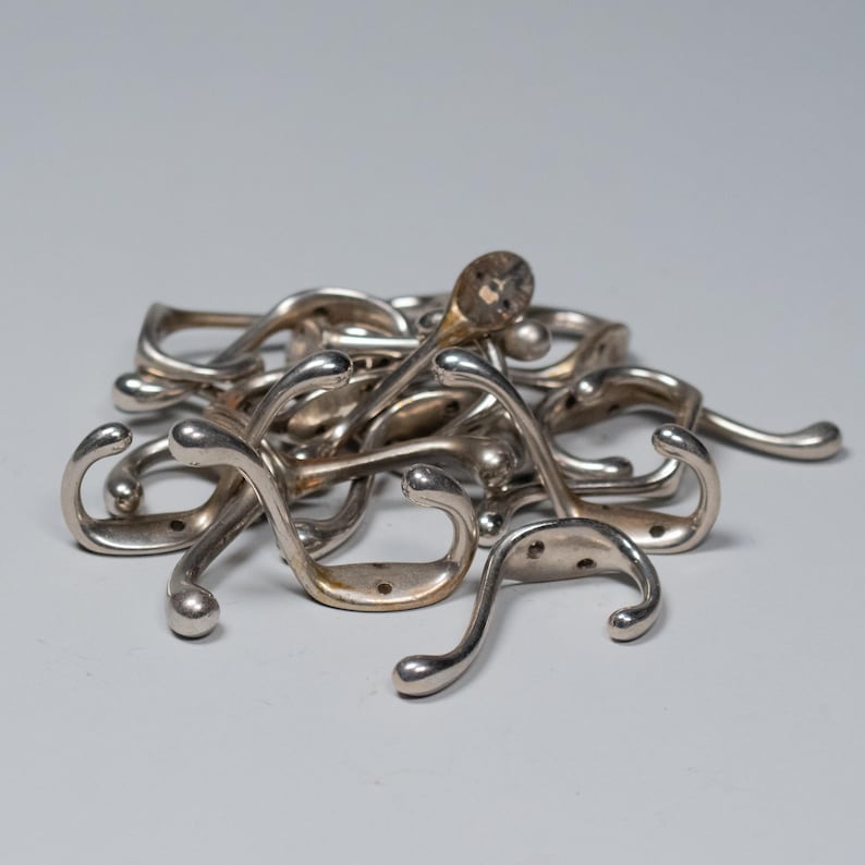 Op de afbeelding: Een stapel zilverkleurige metalen kapstokken. Elke haak heeft een gebogen ontwerp met een afgerond uiteinde en een platte basis met twee gaten voor montage. De haken liggen verspreid op een lichtgrijze ondergrond.