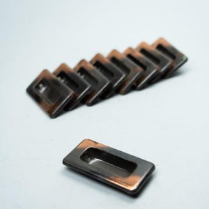 Op de afbeelding: Een verzameling rechthoekige, verzonken kastgrepen met een donkere, metallic afwerking en koperkleurige randen. De grepen zijn diagonaal gerangschikt, met één greep geïsoleerd op de voorgrond.