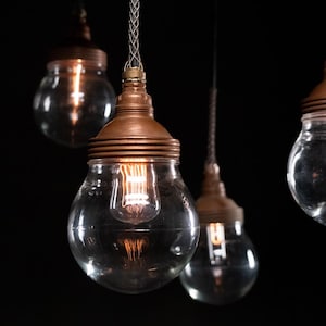 May include: Close-up of several hanging pendant lights. Each light features a clear, round glass bulb with a copper-colored metal top. The bulbs are illuminated, showcasing the warm glow of the filaments. The lights hang against a dark background.