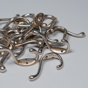 Op de afbeelding: Een verzameling vintage zilverkleurige metalen kapstokhaken. De haken hebben een gebogen ontwerp met een afgerond uiteinde en een platte basis met schroefgaten voor montage. Sommige haken vertonen tekenen van slijtage.