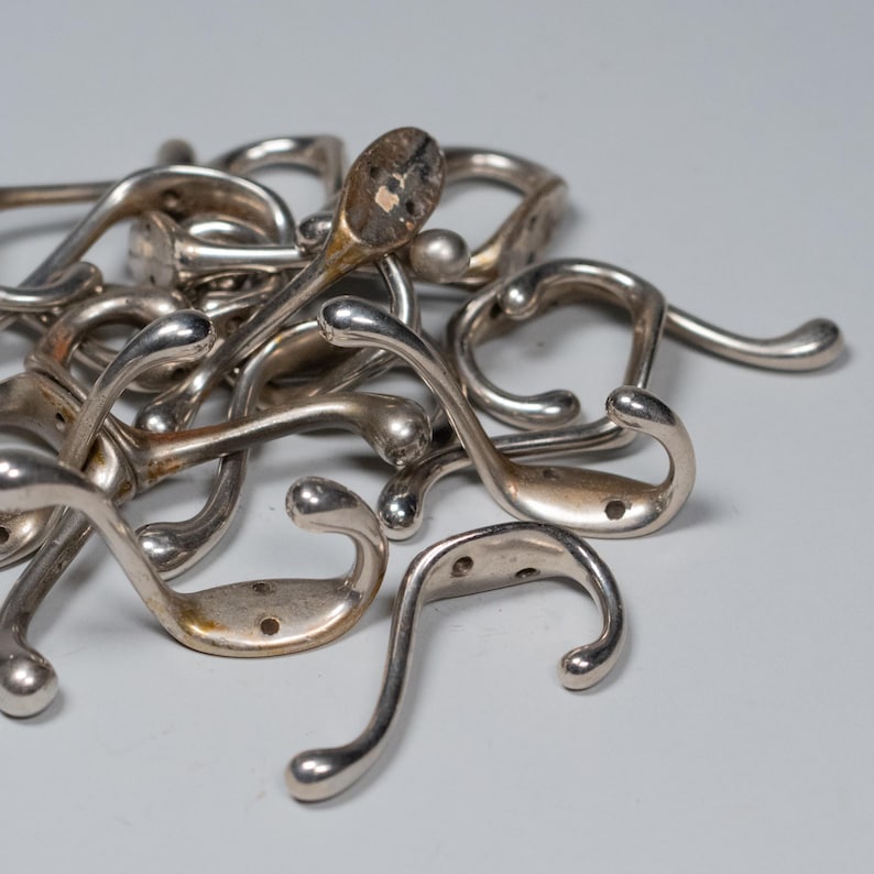 Op de afbeelding: Een verzameling vintage zilverkleurige metalen kapstokhaken. De haken hebben een gebogen ontwerp met een afgerond uiteinde en een platte basis met schroefgaten voor montage. Sommige haken vertonen tekenen van slijtage.