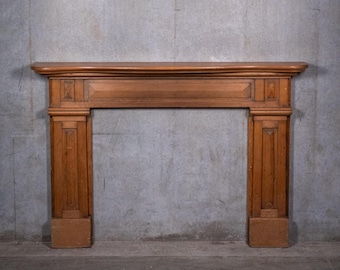May include: A medium-brown wooden fireplace mantel featuring a horizontal shelf, a central rectangular panel, and two vertical side pillars. Each pillar includes a recessed rectangular panel with a small diamond-shaped accent at the top.