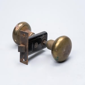 Peut inclure: Ensemble de poignées de porte en laiton antique avec une plaque de verrouillage noire. Les boutons sont ronds et présentent des signes d'usure. La plaque de verrouillage est rectangulaire avec un trou de serrure et des trous de vis.