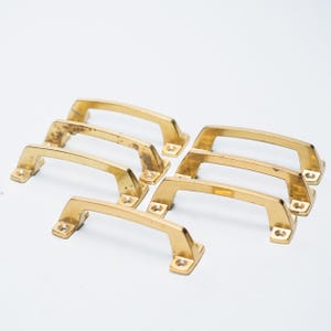 May include: A collection of seven vintage brass cabinet pulls. The pulls have a rectangular base with two screw holes and a curved handle. Some pulls show signs of age with slight discoloration. The hardware is set against a white background.